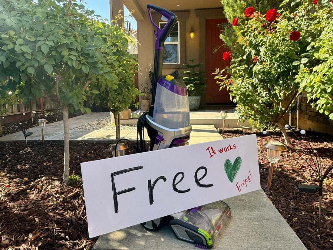 A carpet cleaner on a front lawn beside a hand-lettered sign reading Free — it works, enjoy